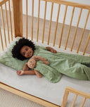Toddler lays in bed, wearing an ergoPouch Sleep Suit Sack in Willow with matching Doll Sack and Pillowcase