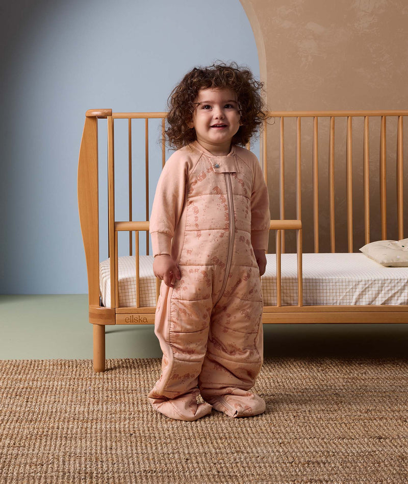A toddler dressed in a 2.5 TOG Sleep Suit Sack stands next to a toddler bed.