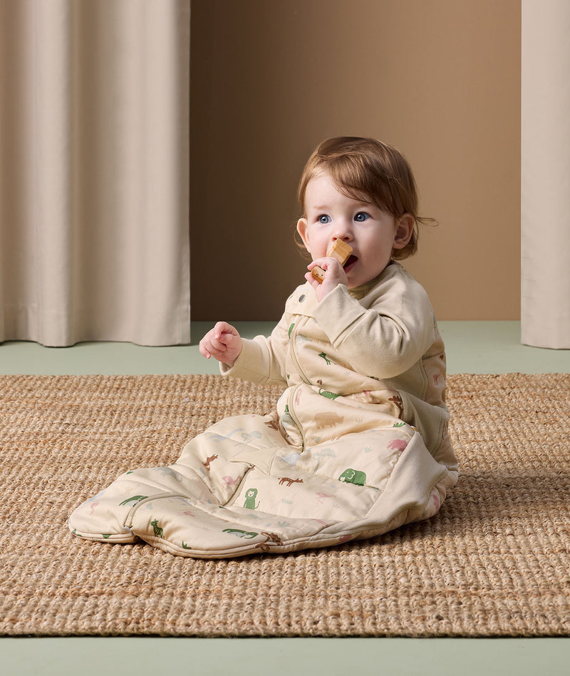 An infant is sitting on a rug, wearing an ergoPouch Sleep Suit Sack and putting a toy in its mouth.