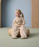 A little girl sits on a woven cushion wearing an ergoPouch sleep suit sack with long sleeves and a front zipper. The suit features a cozy, padded design with colorful animal prints.