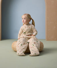A little girl sits on a woven cushion wearing an ergoPouch sleep suit sack with long sleeves and a front zipper. The suit features a cozy, padded design with colorful animal prints.