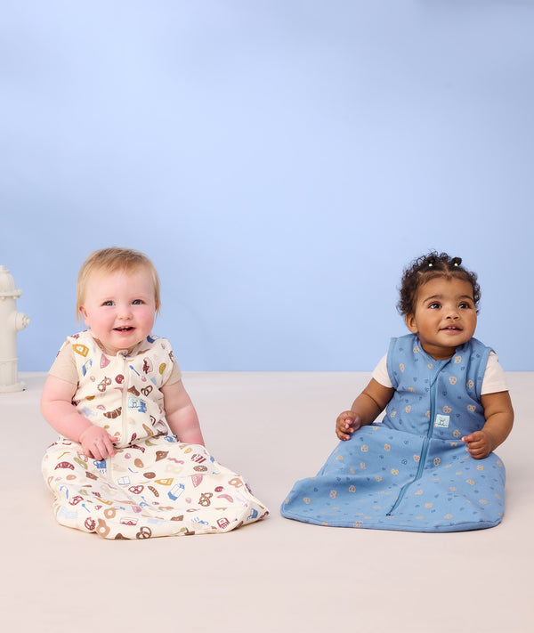 Two infants sitting on the floor wearing ergoPouch Sleep Sack 0.2 TOG in Bon Appetit and Petite Pretzel