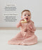 A baby sitting on a cream rug, holding a wooden ring toy to its mouth while wearing a pink floral sleep sack with text highlighting organic cotton and stretchy & bell-shaped design.