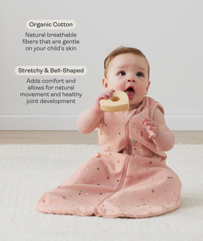 A baby sitting on a cream rug, holding a wooden ring toy to its mouth while wearing a pink floral sleep sack with text highlighting organic cotton and stretchy & bell-shaped design.