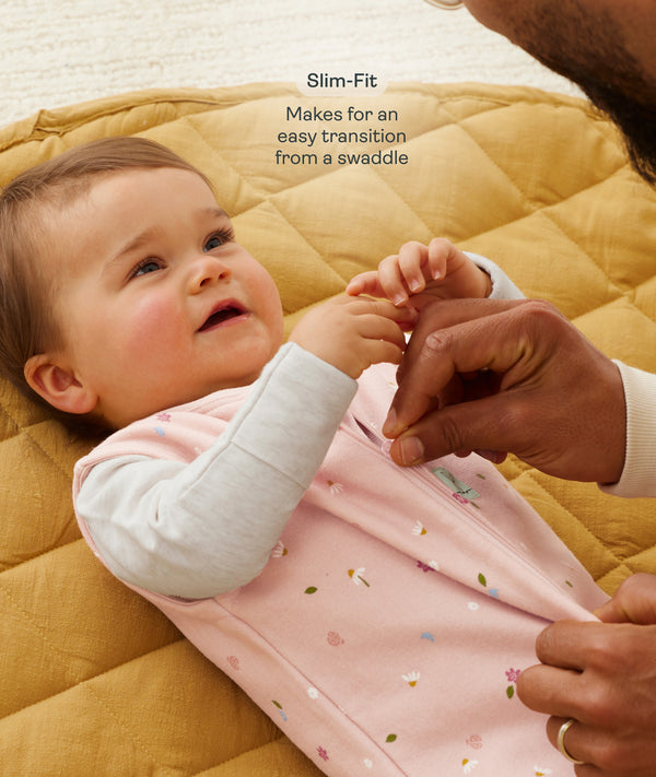 A close-up view of a baby lying on a mustard-yellow quilted mat while an adult gently adjusts the zipper of the baby's pink floral sleep sack with text overlay highlighting slim-fit design for an easy transition from swaddling.