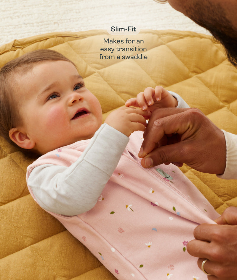 A close-up view of a baby lying on a mustard-yellow quilted mat while an adult gently adjusts the zipper of the baby's pink floral sleep sack with text overlay highlighting slim-fit design for an easy transition from swaddling.