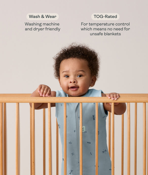 Infant wearing a sleeveless, light blue dragonfly-patterned sleep sack stands in a wooden crib, holding onto the top rail with both hands. Above the baby, text features highlights for the product: Wash & Wear and TOG-Rated.