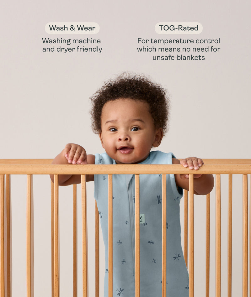 Infant wearing a sleeveless, light blue dragonfly-patterned sleep sack stands in a wooden crib, holding onto the top rail with both hands. Above the baby, text features highlights for the product: Wash & Wear and TOG-Rated.