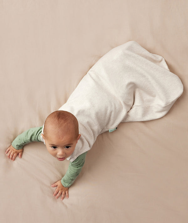 Baby crawling on a plain surface while wearing an oatmeal-colored sleep sack over a green long sleeved romper.