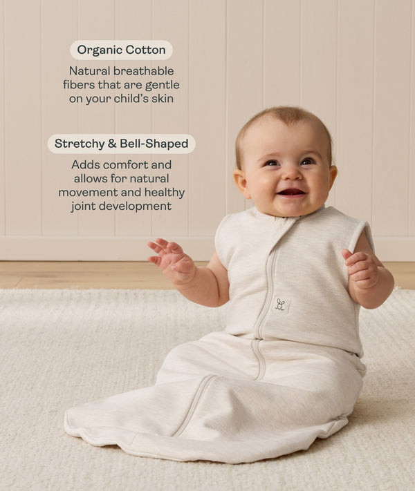 Smiling infant sitting on a white mat, wearing a sleeveless Sleep Sack in Oatmeal Marle, with text highlighting organic cotton and stretchy, bell-shaped design.