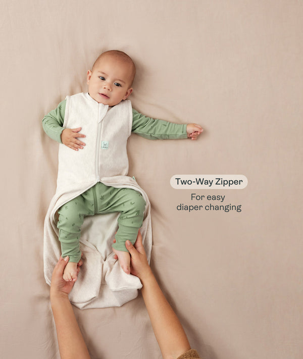 A baby lying down in a green long sleeve romper with an unzipped oatmeal-colored sleep sack while adult hands hold their feet. Text highlighting 