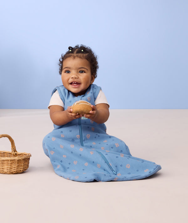 Infant sitting on the floor wearing ergoPouch Sleep Sack 0.2 TOG in Petit Pretzel and holding a fake pretzel toy