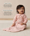 Infant sitting upright on a white rug wearing a pink long sleeve Sleep Sack  with text highlighting organic cotton and stretchy & bell-shaped design.