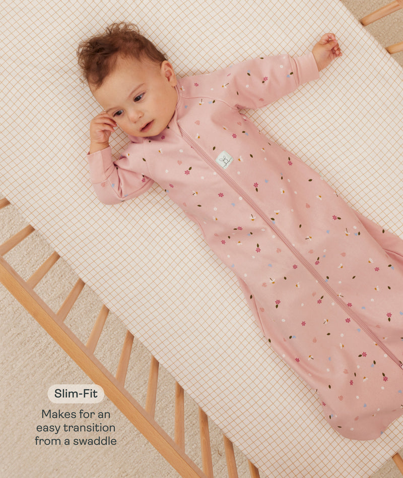 Infant lying in a wooden crib wearing a pink floral sleeved sleep sack with text highlighting slim fit design for an easy transition from swaddling.
