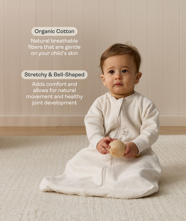 Infant sitting, wearing a sleeved Sleep Sack 1.0 TOG Oatmeal Marle, while holding a wooden toy. Text highlighting organic cotton and stretchy-bell-shaped design.
