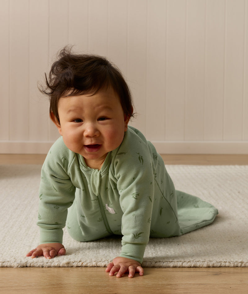 A baby wearing a long-sleeved, sage green leaf-patterned sleep sack crawls on a light rug in front of a neutral wood-paneled wall, smiling toward the camera.