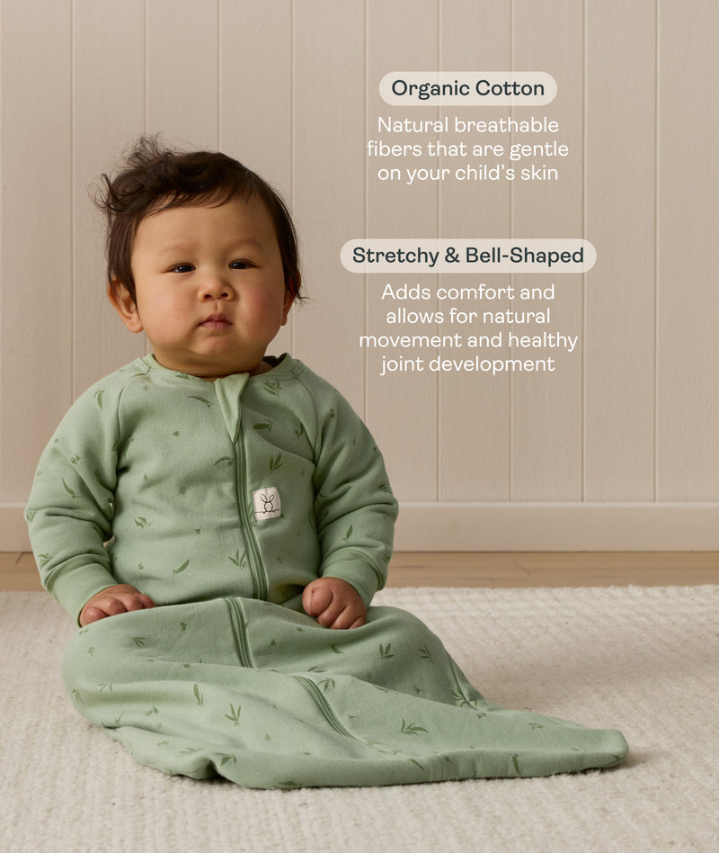 Infant sitting on a white mat, wearing a Sleeved Sleep Sack 1.0 TOG Willow with text overlay highlighting organic cotton and stretchy & bell-shaped design.