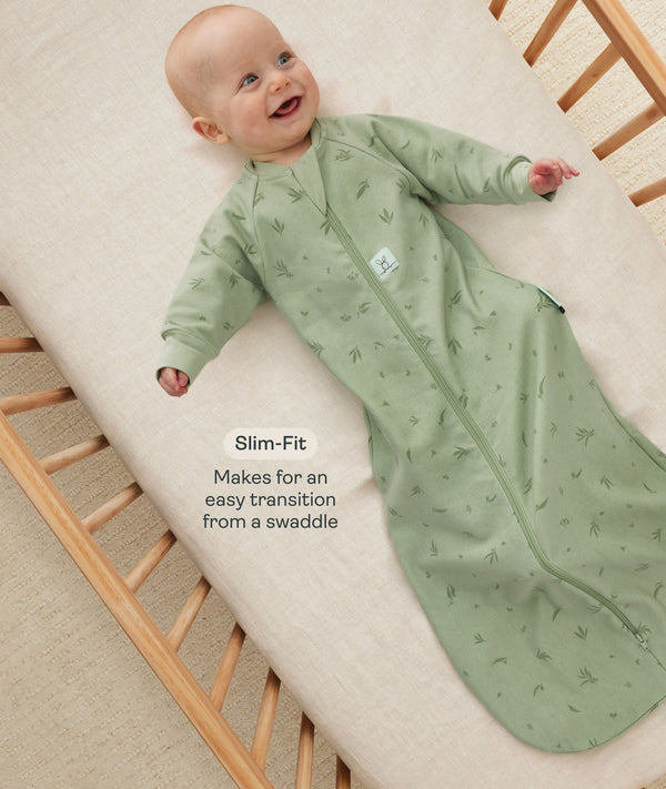 Happy baby lying in a wooden crib wearing a green long-sleeved sleep sack with text overlay highlighting slim-fit design for an easy transition from swaddling.