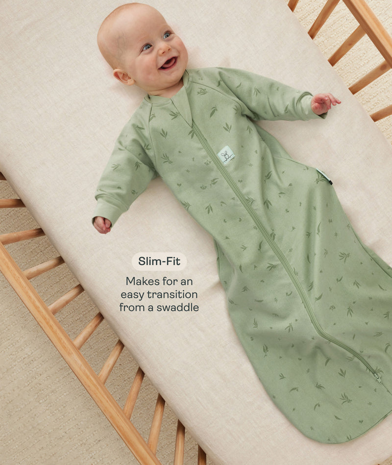 Happy baby lying in a wooden crib wearing a green long-sleeved sleep sack with text overlay highlighting slim-fit design for an easy transition from swaddling.