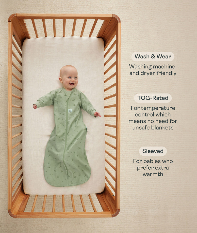 An overhead view of a baby wearing a long-sleeved, sage green leaf-patterned sleep sack lying on their back inside a wooden crib. To the right, text details key features: wash & wear, TOG-rated, and Sleeved.