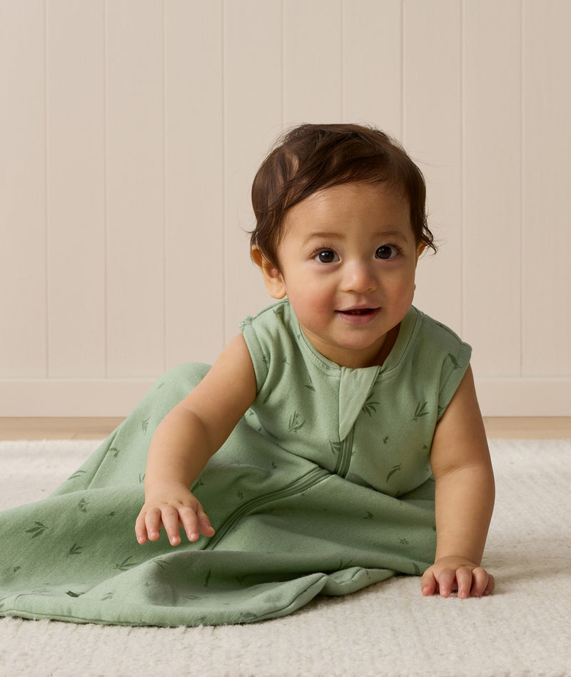 A baby wearing a sleeveless, sage green leaf-patterned sleep sack sits on a light rug, leaning forward with one hand resting on the floor.
