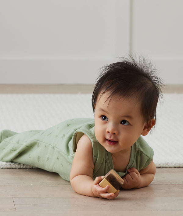 A baby wearing a sleeveless, sage green leaf-patterned sleep sack lies on their stomach on a wooden floor, holding a small wooden block and looking off to the side.