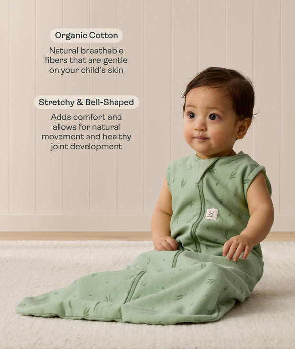 Infant sitting on a white mat, wearing a green Sleep Sack 1.0 TOG with text highlighting organic cotton and stretchy & bell-shaped design.