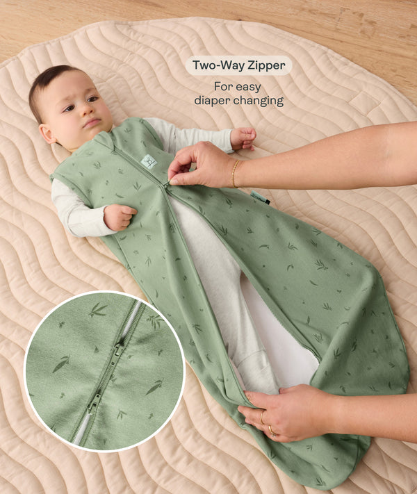 Baby lying on a padded mat while an adult adjusts the zipper of the baby's green sleeveless sleep sack with text overlay highlighting two-way zip design for easy diaper changing.