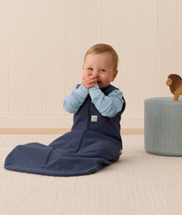 Smiling infant in an ergoPouch Sleep Sack 1.0 TOG Ink sitting on a light wooden floor with a neutral wall background.
