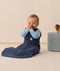 Smiling infant in an ergoPouch Sleep Sack 1.0 TOG Ink sitting on a light wooden floor with a neutral wall background.