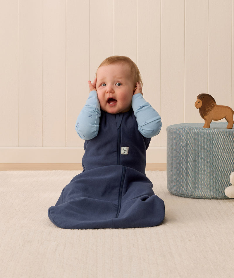 Infant in an ergoPouch Sleep Sack 1.0 TOG Ink sitting on a light wooden floor with a neutral wall background.