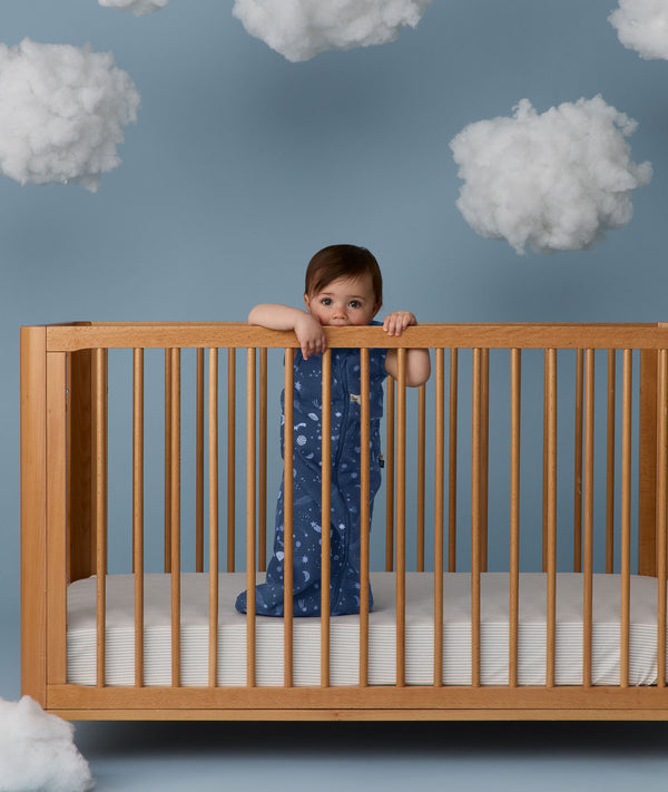 Child standing in a wooden crib wearing an ergoPouch Sleep Sack 1.0 TOG Starbeam
