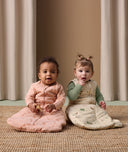 Two babies sitting side by side wearing ergoPouch Sleep Sack 3.5 TOG Sleeved in Elephant Parade and Sleep Sack 2.5 TOG Savannah