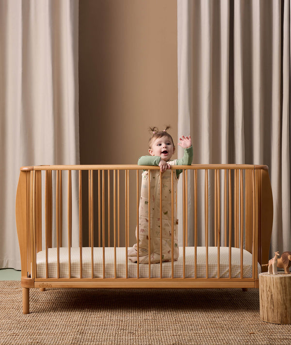 A baby standing in a wooden crib wearing ergoPouch Sleep Sack with cute animal prints