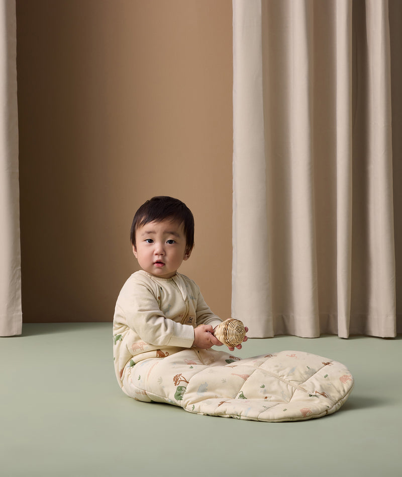 Infant wearing ergoPouch Sleep Sack with Sleeves in Savannah print, sitting on the floor while holding a toy.
