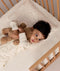 Toddler lying in a wooden crib on a cream patterned Organic Toddler Pillow while hugging a stuffed teddy bear.