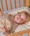 A toddler lies on their back in a wooden crib, resting their head on a patterned cream Organic Toddler Pillow while wearing a beige printed sleep suit.