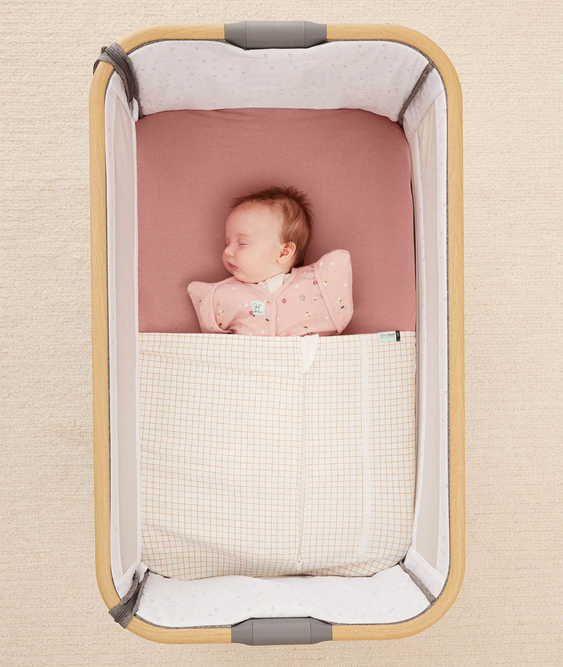 Newborn baby asleep in a bedside sleeper under a ergoPouch HugMe Blanket in Caramel Grid