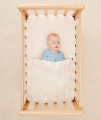 Newborn baby asleep in a bassinet under a ergoPouch HugMe Blanket in Caramel Grid