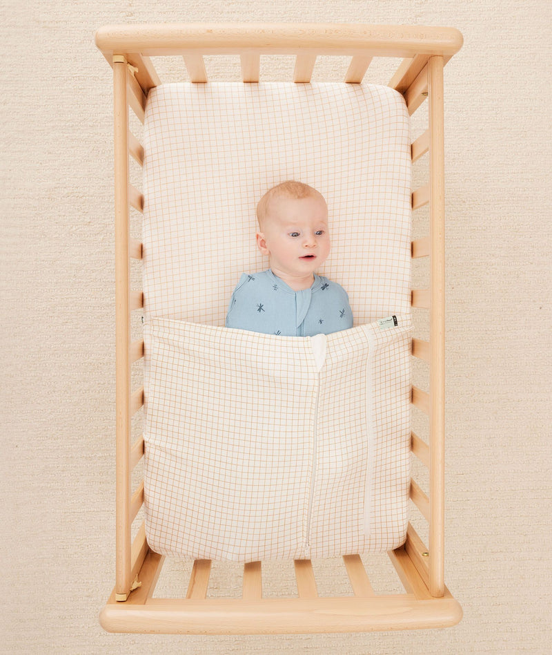 Newborn baby asleep in a bassinet under a ergoPouch HugMe Blanket in Caramel Grid