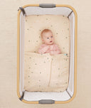 Baby lying in a bedside sleeper under an ergoPouch HugMe Blanket in Daisy Sprinkle