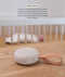 A compact, circular Drift Away white noise machine with a carry strap rests on a wooden surface. In the softly blurred background, a baby sleeps safely in a crib. Text overlay titled 