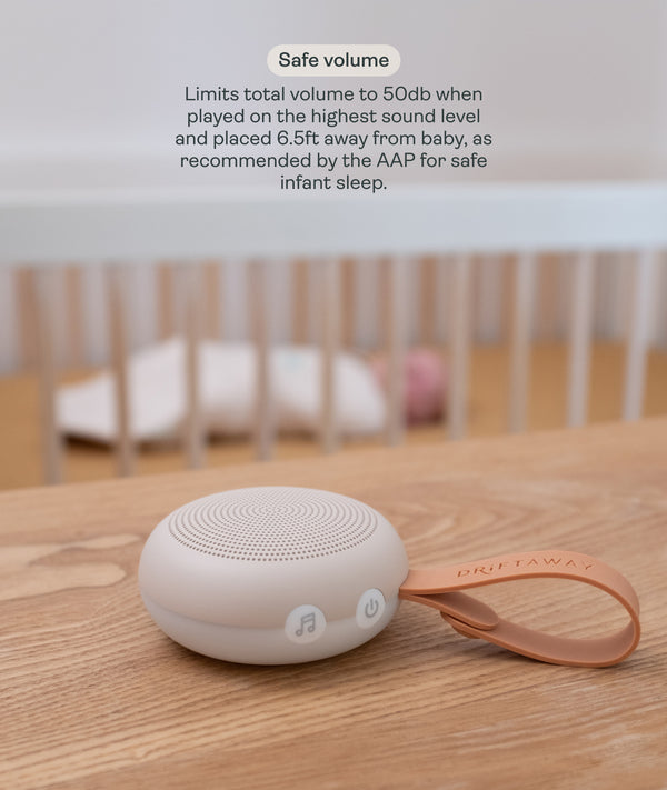A compact, circular Drift Away white noise machine with a carry strap rests on a wooden surface. In the softly blurred background, a baby sleeps safely in a crib. Text overlay titled 