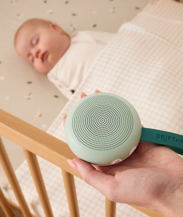 A parent holding a sage color portable white noise machine with a dark green strap, positioned next to a sleeping baby in a wooden crib.
