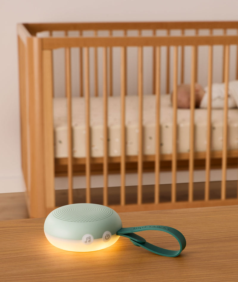 A soft green white noise machine with a glowing warm night light sits on a wooden table in the foreground. It features buttons, including music and power icons, and a green strap with the brand name “Drift Away.” In the background, a baby is peacefully sleeping in a wooden crib, emphasizing the calming effect of the device.