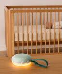 A portable white noise machine with a glowing night light and dark green strap is placed on a wooden surface in the foreground, with a sleeping baby in a wooden crib visible in the background.