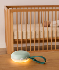 A portable white noise machine with a glowing night light and dark green strap is placed on a wooden surface in the foreground, with a sleeping baby in a wooden crib visible in the background.