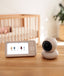 ergoPouch Drift Home Baby Monitor sits on a wooden surface in a softly lit nursery. The setup includes a white video monitor screen displaying sleepwear recommendations with temperature-related TOG ratings, and a matching white camera unit with a black lens. In the blurred background, a baby sleeps peacefully in a wooden crib, emphasizing a calm and safe sleep environment.