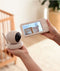A parent holds a baby monitor system, consisting of a white camera unit with a black lens in one hand and a handheld video monitor in the other. The monitor screen shows a live video feed of a wooden crib in a softly lit nursery, highlighting the product’s real-time monitoring capability and clear visual display.