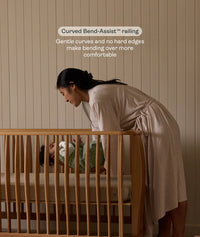 Woman placing baby into the ergoPouch ellska Skye Crib, supported by ergonomic Bend-Assist™ railings for easier access